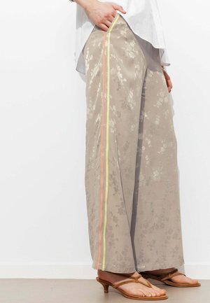 Pantalon large beige avec un motif floral subtil et des bandes latérales jaunes et oranges, porté avec des sandales à talons marron clair et un haut blanc.