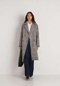 Διπλής όψης γκρι καρώ παλτό από μάλλινο ύφασμα με αναγνωρίσιμα πέτα, μπροστινές τσέπες και άνετη εφαρμογή, που φοριέται πάνω από ένα λευκό πουκάμισο και σκούρους παντελόνες.