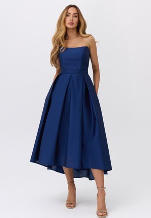 Kobieta o długich falowanych włosach, ubrana w granatową sukienkę midi bez ramiączek i beżowe sandały na kostkę, stojąca na jednolitym tle.