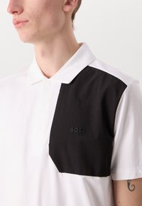 Balta trumpas rankovėmis polo marškinėliai su didele juoda tekstūruota juosta kairėje, su iškilniu "BOSS" logotipu krūtinėje.