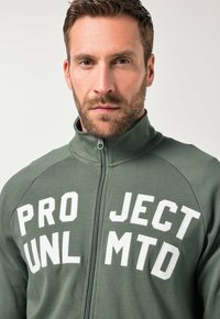 Giacca verde con zip e testo ricamato bianco "PRO JECT UNL MTD." Presenta un colletto alto e un design aderente. Tessuto dalla consistenza morbida.