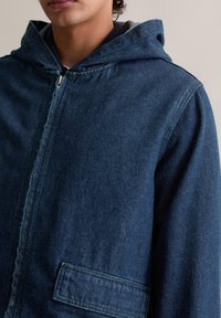 Dunkelblaue Jeansjacke mit Kapuze, mit einem Reißverschluss vorne, einer seitlichen Tasche und einer strukturierten Stoffoberfläche.
