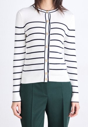 Vrouw draagt een witte geribbelde cardigan met marineblauwe horizontale strepen en gouden knopen, gecombineerd met donkergroene broek.