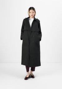 Una donna è in piedi contro uno sfondo bianco, indossando un lungo trench coat nero, una maglietta bianca, pantaloni cropped scuri e scarpe bassi in vernice bordeaux lucide.