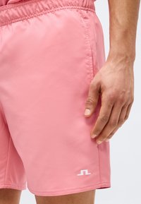 Άνδρας που φοράει ροζ σορτς με ελαστική μέση και μικρό λευκό λογότυπο κοντά στο τελείωμα, με το χέρι χαλαρό δίπλα στο σώμα.