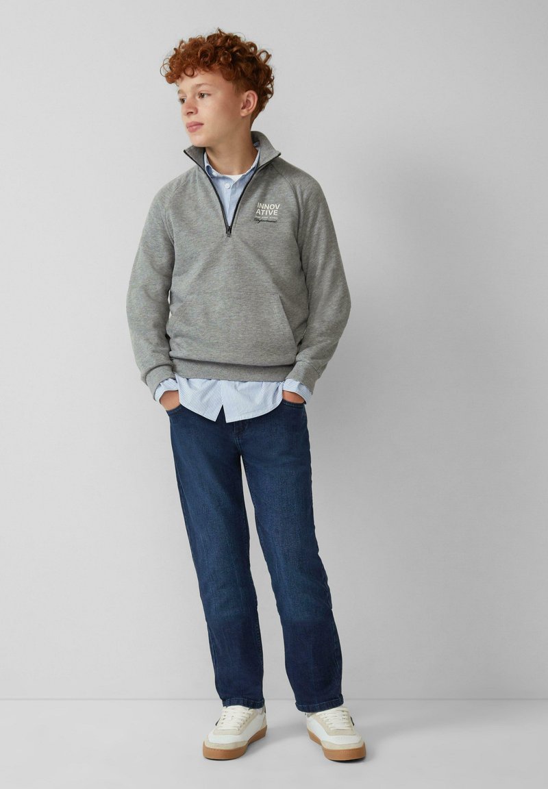Grijze rits sweatshirt met geribde manchetten, over een lichtblauw gestreept shirt, gecombineerd met donkerblauwe jeans en witte sneakers.