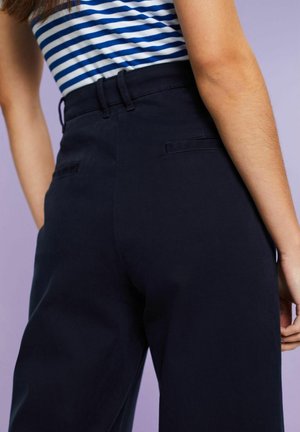Granatowe spodnie o wysokim stanie z gładką fakturą, z bocznymi kieszeniami i szlufkami na pasek. Widoczne detale szwu z tyłu. Dopasowane w biodrach, szerokie nogawki.