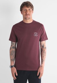 Un homme aux cheveux bruns courts et avec des tatouages sur les deux bras porte un T-shirt bordeaux avec un graphisme blanc "Oxbow 1985" sur la poitrine gauche.