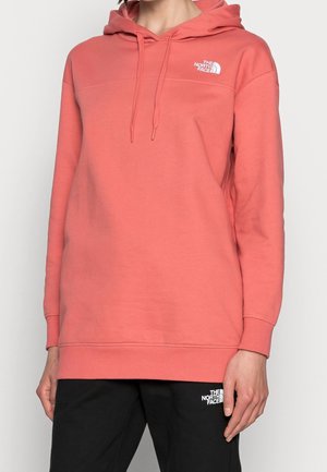 Person klädd i en korallfärgad The North Face-huvtröja med logotyp på bröstet och svarta byxor med logotyp på låret, som står framför en enfärgad bakgrund.