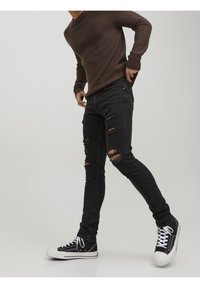 Jeans skinny desgastados negros con desgarros prominentes, combinados con zapatillas altas negras y un suéter marrón de canalé, sobre un fondo liso.