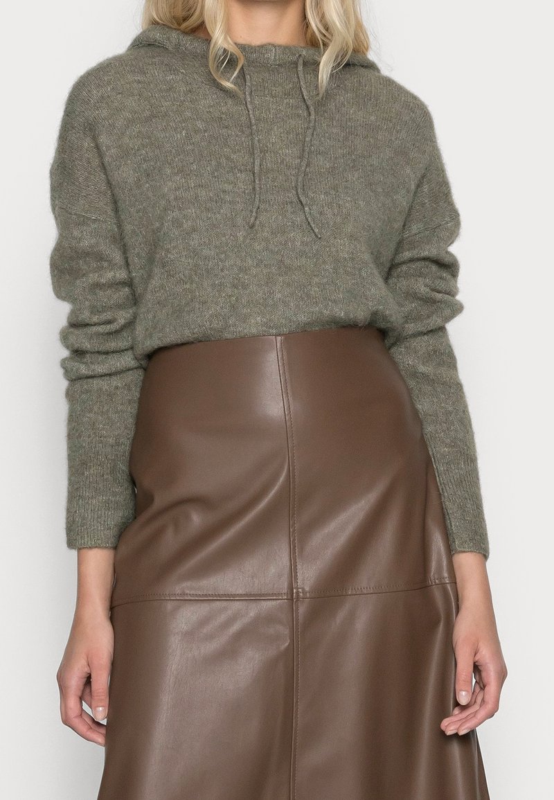 Femme portant un pull à capuche en maille vert olive rentré dans une jupe en cuir marron taille haute, debout devant un fond uni.