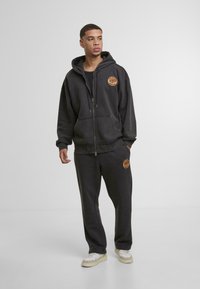 Sötét szürke kapucnis pulóver és melegítőnadrág szett, puha anyagból készült, elöl cipzárral, nagy kapucnival és narancssárga logóval a mellrészen.