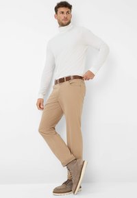 Beige Baumwollhose, taillierter Schnitt, kombiniert mit einem weißen Rollkragenoberteil. Brauner Ledergürtel, graue Schnürstiefel mit Gummisohlen.