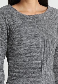 Robe grise texturée avec un large décolleté, présentant des détails en panneaux contrastants. Le tissu est doux avec des rayures horizontales et verticales.