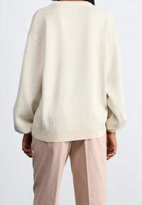 Pull oversize couleur crème, en maille texturée, épaules tombantes et ourlet côtelé, porté avec un pantalon rose clair.