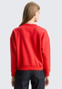 Rode trui met een losse pasvorm, ronde halslijn en geribbelde manchetten, gecombineerd met donkere denim jeans. Zacht katoenen materiaal zichtbaar.