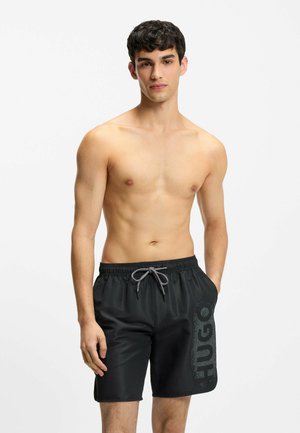 Młody mężczyzna stojący bez koszulki, ubrany w czarne kąpielówki z logo "HUGO" na lewej nogawce, ręce opuszczone wzdłuż ciała.