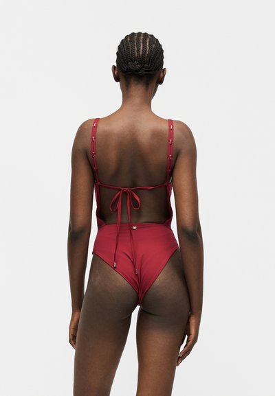 Γυναίκα με πλεκτό μαλλί που φοράει ένα μαρτζανά κόκκινο ολόσωμο μαγιό με ρυθμιζόμενους ιμάντες και δεμένη λεπτομέρεια στην πλάτη, μπροστά σε απλό φωτεινό φόντο.