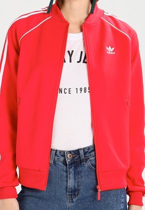 Veste zippée rouge Adidas avec des bandes blanches sur les manches portée sur un t-shirt blanc imprimé et un jean bleu.