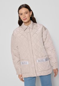 Beige gewatteerde jas met oversized pasvorm, drukknoopsluitingen en twee voorzakken met witte merkaccenten. Gladde, glanzende textuur.