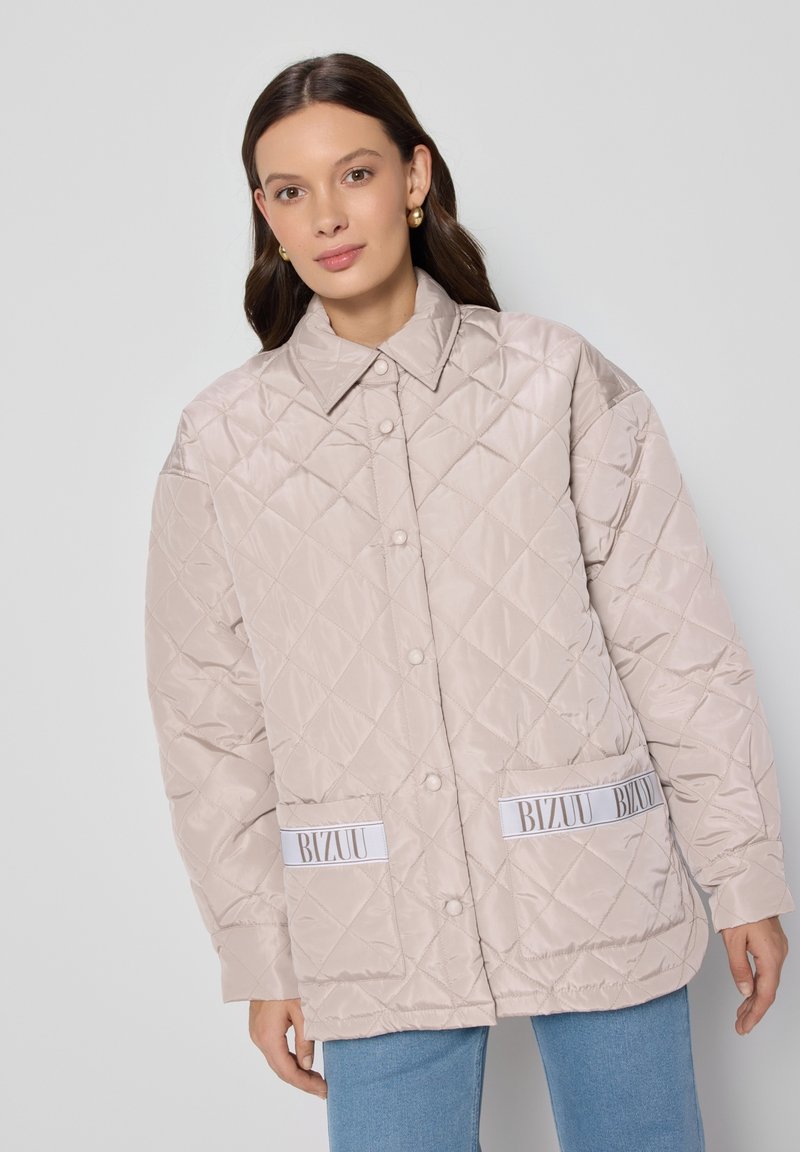 Beige gewatteerde jas met oversized pasvorm, drukknoopsluitingen en twee voorzakken met witte merkaccenten. Gladde, glanzende textuur.