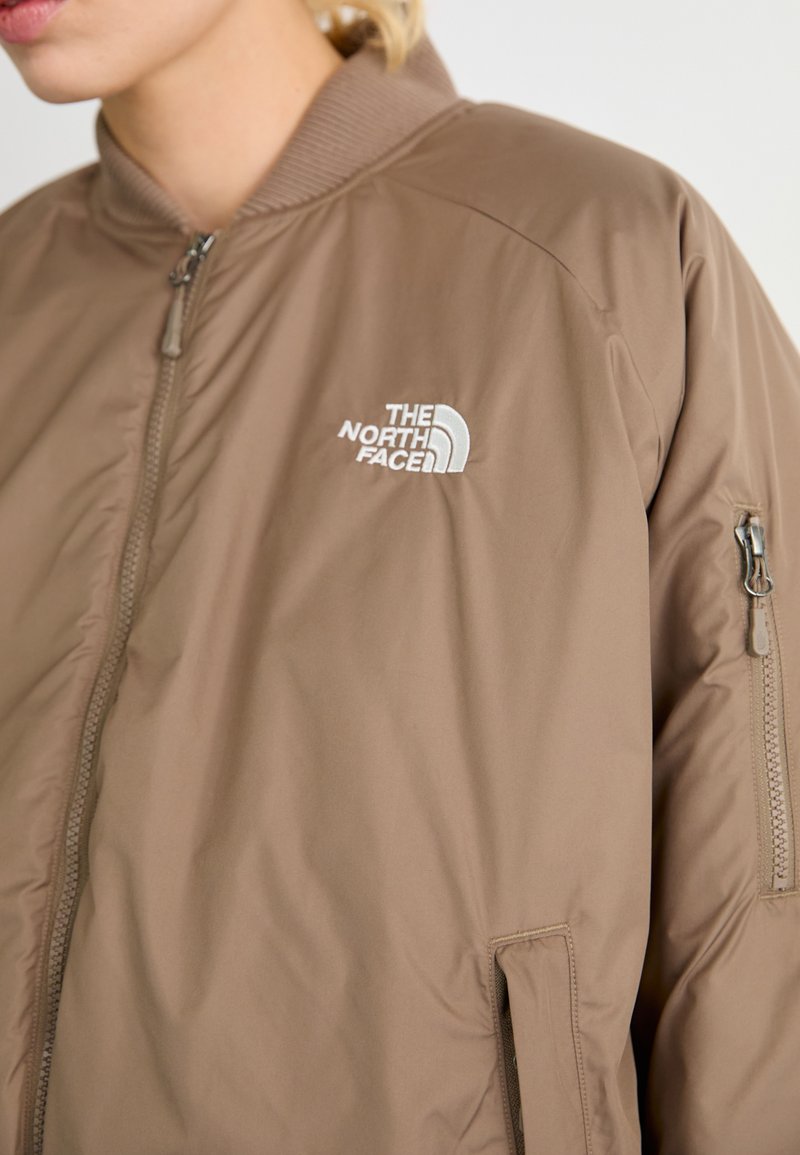 Giubbotto bomber imbottito marrone con colletto a costine e tasca laterale con zip, dotato di una patch con logo bianco sul petto sinistro. Superficie liscia.