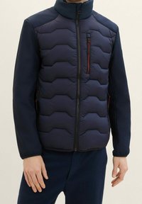 Mörkblå pufferjacka med texturerade paneler, med hög krage, dragkedjefick på bröstet och stickade ärmar. Hexagonal kviltad mönster.