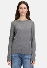 Eine Frau mit langen braunen Haaren, die einen grauen Strickpullover und blaue Jeans trägt, steht vor einem schlichten weißen Hintergrund.