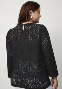 Zwarte blouse met lange mouwen, voorzien van een uitsnijdingspatroon en een sleutelgatdetail op de rug. Zachte stof met textuele designelementen.