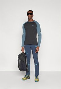 Zwart shirt met lange mouwen en blauwe accenten, gecombineerd met blauwe broek en groene wandelschoenen; model houdt een zwarte rugzak vast.