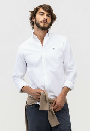Hombre con camisa blanca abotonada con las mangas remangadas, atando un suéter beige alrededor de su cintura, de pie frente a un fondo liso.