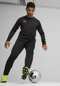 Schwarzes, sportliches Langarmoberteil und Hose mit Puma-Logo; trägt leuchtend gelbe Fußballschuhe und dribbelt einen silbernen Fußball.