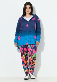 Sudadera azul con patrones de caras sonrientes rosas, cierre de cremallera y cordones. Pantalones de patrón abstracto coloridos en rosa, verde y negro.