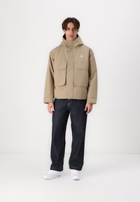 Beige waterproof jacket with a hood, two large front pockets, and a triangular logo. Paired with dark denim jeans and white sneakers.