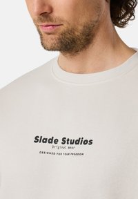 Felpa beige chiaro con collo rotondo, caratterizzata da un logo nero "Slade Studios Original Wear" e la scritta "DESIGNATA PER LA TUA LIBERTÀ."