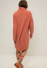Abito a maglia color ruggine con colletto alto, maniche lunghe e vestibilità rilassata, abbinato a stivali alla caviglia marroni e calze a righe.