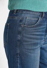 Jeans en denim d'un bleu moyen, avec une coupe ajustée, un design à cinq poches et des coutures contrastantes le long des coutures.