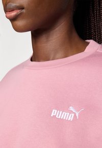 Felpa di colore rosa chiaro con scollatura rotonda; presenta un logo Puma bianco sul petto. Il materiale sembra morbido con una texture liscia.