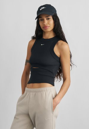 Femme portant une casquette noire Nike et un crop top sans manches avec un pantalon de survêtement beige Nike, debout avec une main dans la poche devant un fond uni.