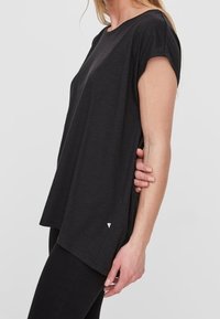 Svart kortärmad topp i mjukt tyg, med avslappnad passform, rund halsringning och en liten triangulär logga vid fållen.