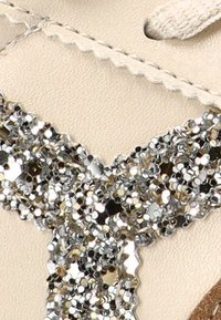 Zapato beige con una brillante tira de lentejuelas plateadas, que presenta una textura suave, puntera redonda y diseño con cordones.