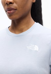 Κοντινό πλάνο ενός ατόμου που φορά ένα ανοιχτό μπλε T-shirt με λευκό λογότυπο "The North Face" στο αριστερό στήθος.