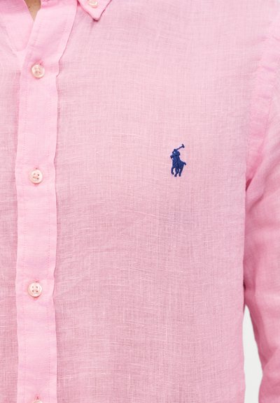 Tuva fotogrāfija ar rozā pogājamu kreklu, uz krūtīm ir neliels tumši zils izšūts polo spēlētāja logotips.