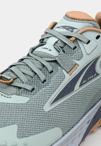 Chaussure de course grise avec tige en mesh, superpositions texturées et accents orange. Les lacets sont plats, avec un embout renforcé et une semelle rembourrée.