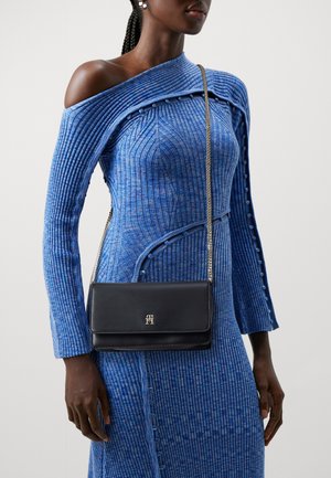 Donna che indossa un vestito blu a coste con una spalla, adornato da dettagli con bottoni, mentre porta una borsa a tracolla in pelle nera con una catena dorata.