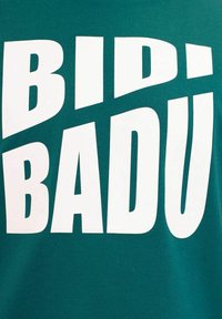 Camiseta verde con un gran texto blanco que dice "BIDI BADU", utilizando una fuente sans-serif en negrita con tamaños y ángulos de letras variados.