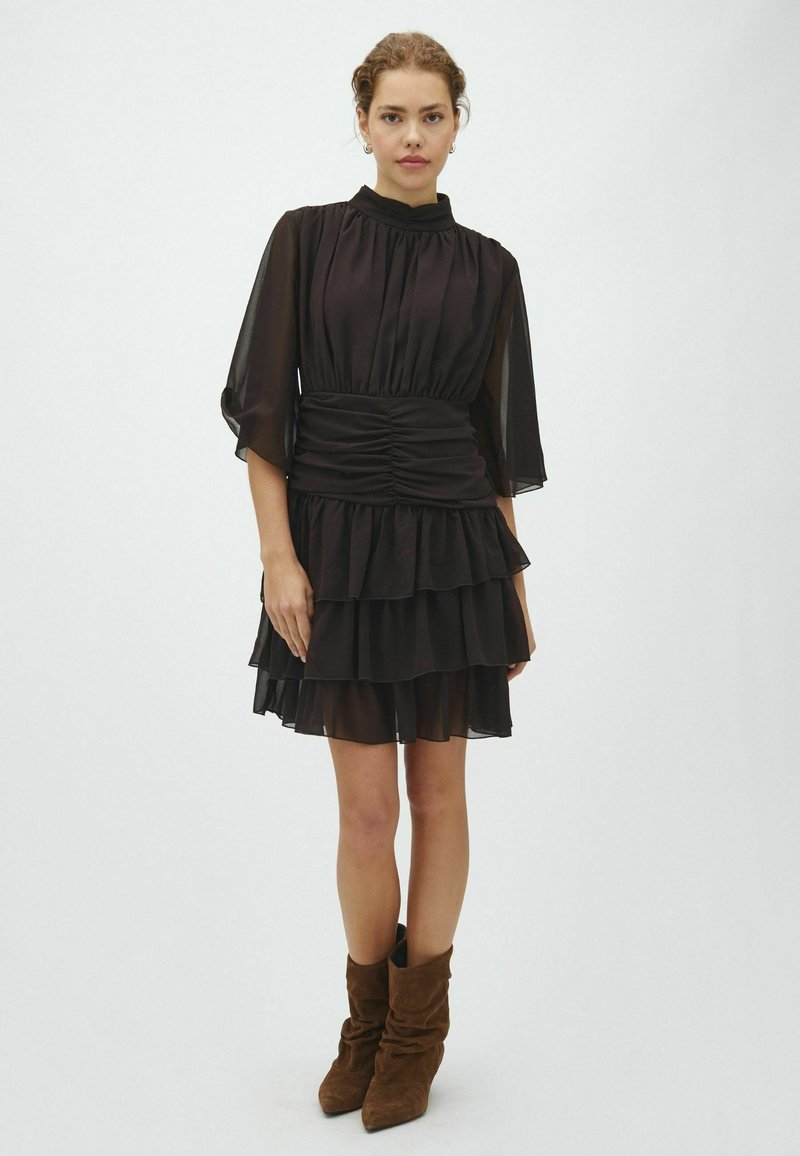 Femme aux cheveux bouclés portant une robe noire transparente à étages avec une taille plissée et des bottes souples en daim marron, debout devant un fond clair uni.