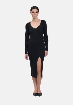 Donna che indossa un abito midi nero aderente a coste con maniche lunghe, scollo a cuore e spacco anteriore alto sulla coscia, in piedi rivolta in avanti.