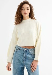 Suéter recortado de punto acanalado crema con cuello alto y mangas largas, combinado con jeans de mezclilla de talle alto en un tono azul claro.