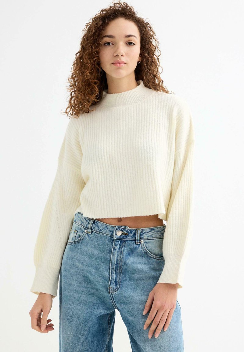 Suéter recortado de punto acanalado crema con cuello alto y mangas largas, combinado con jeans de mezclilla de talle alto en un tono azul claro.